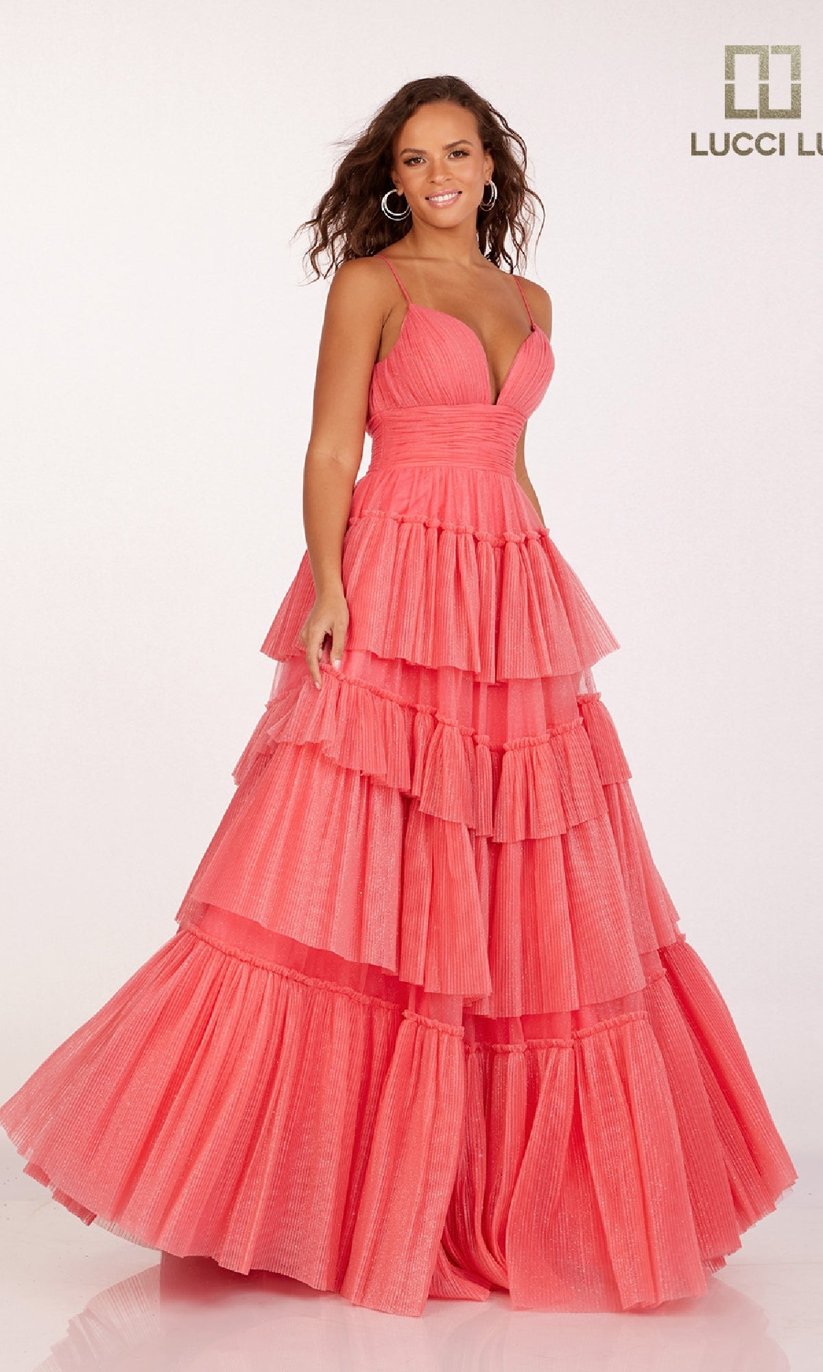 A woman smiles against a plain background with the Lucci Lu logo, wearing the Tiered Ruffled Glitter Prom Dress: Lucci Lu 1357—a sleeveless, floor-length coral pink gown with a deep V-neck and tiered ruffled layers.